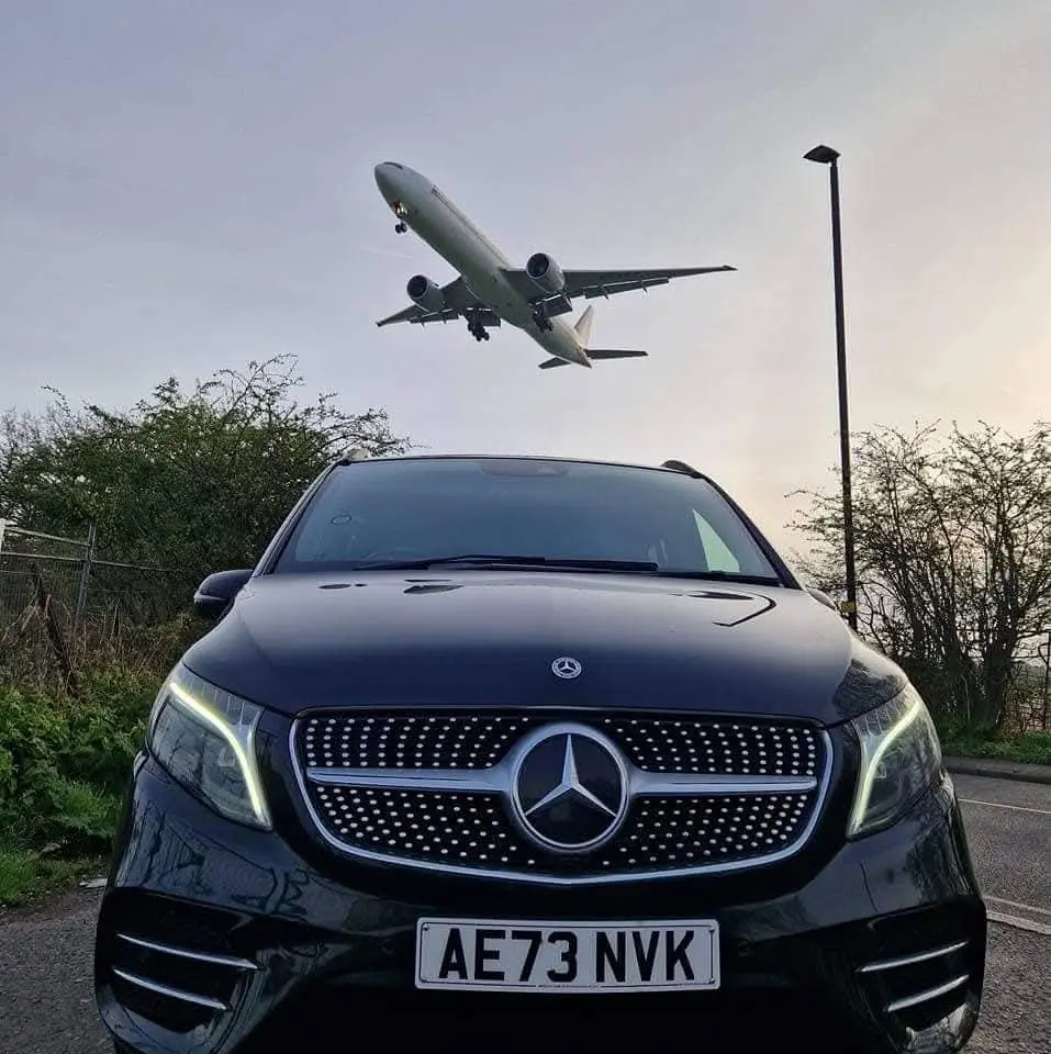 A black Mercedes V-Class with a plane coming in to land overhead